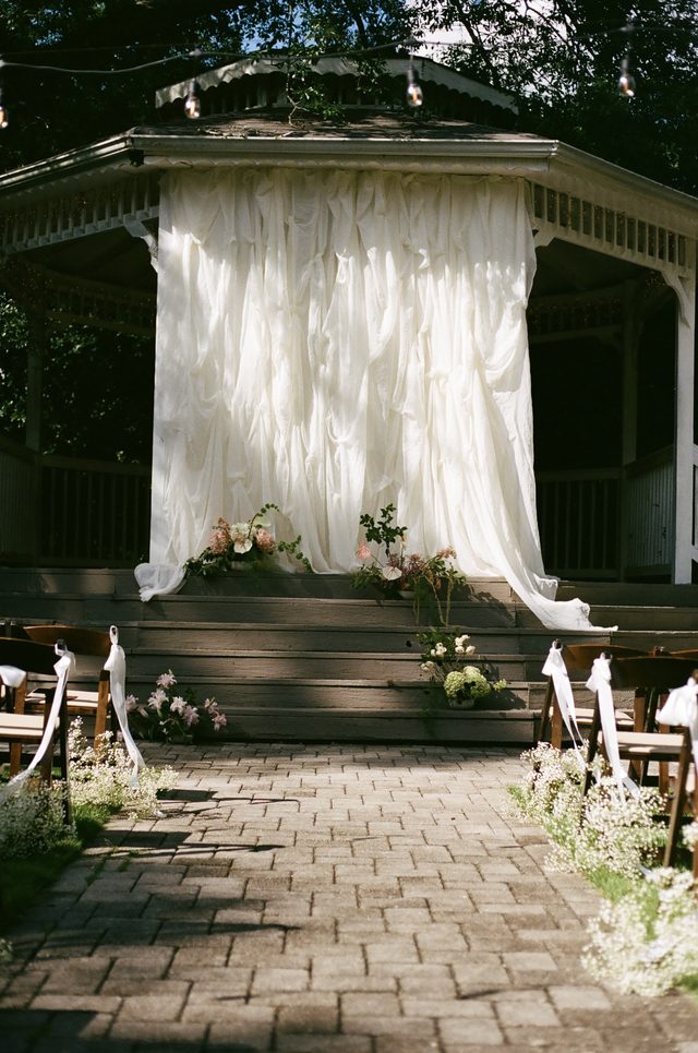 A garden gazebo draped in ivory fabric and flowers at the end of a brick aisle.