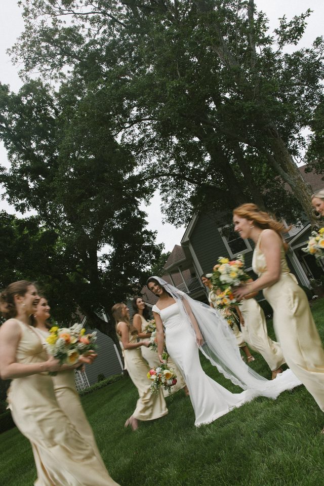 A bride running across a lawn with her bridesmaids, holding yellow bouquets.