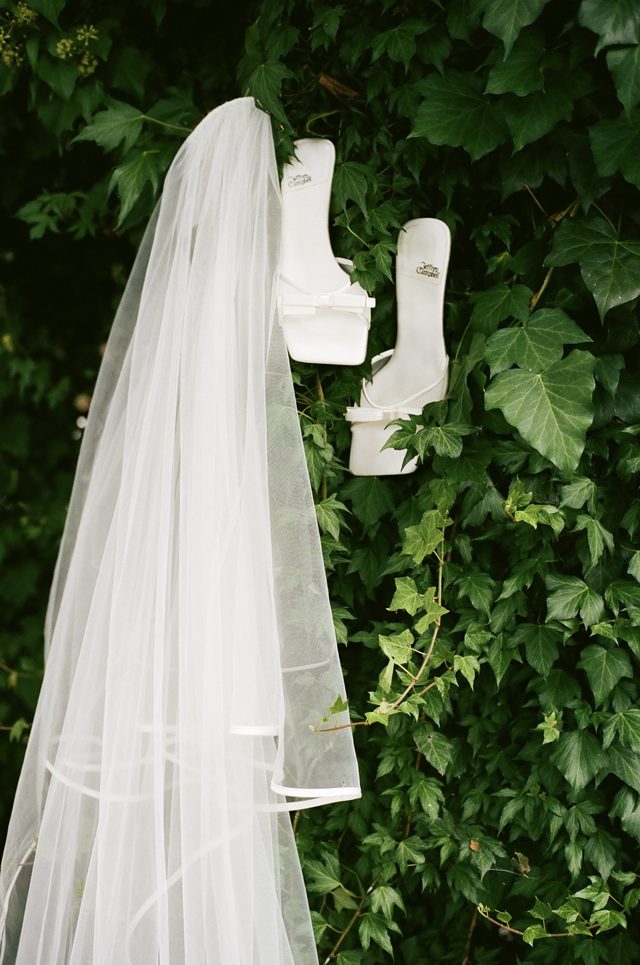 A veil and white heels hanging against a wall of green ivy.