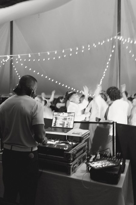The DJ at his booth with the dance floor filling up behind him, in black and white.