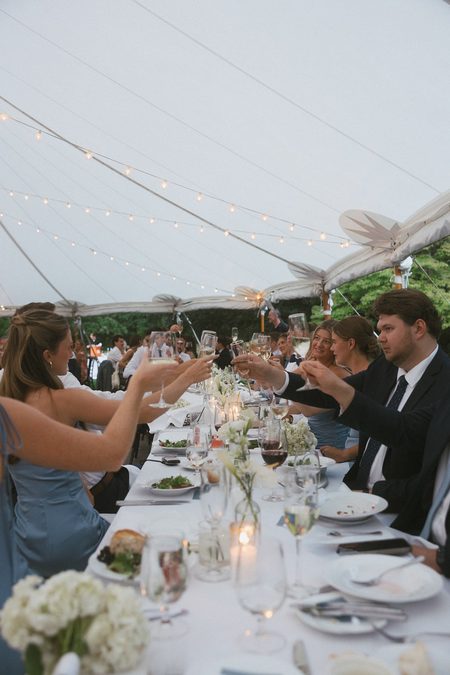 Guests raising their glasses down the long table under the tent, strung with lights.